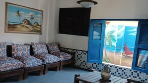 Una habitación con techo de madera, un cuadro de una playa, un conjunto de sofás con cojines estampados, piso de baldosas y vista a través de una ventana con contraventanas.