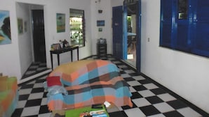 Una habitación con piso a cuadros, cama con manta de colores, ventana con contraventanas azules y puerta que da a otra habitación.