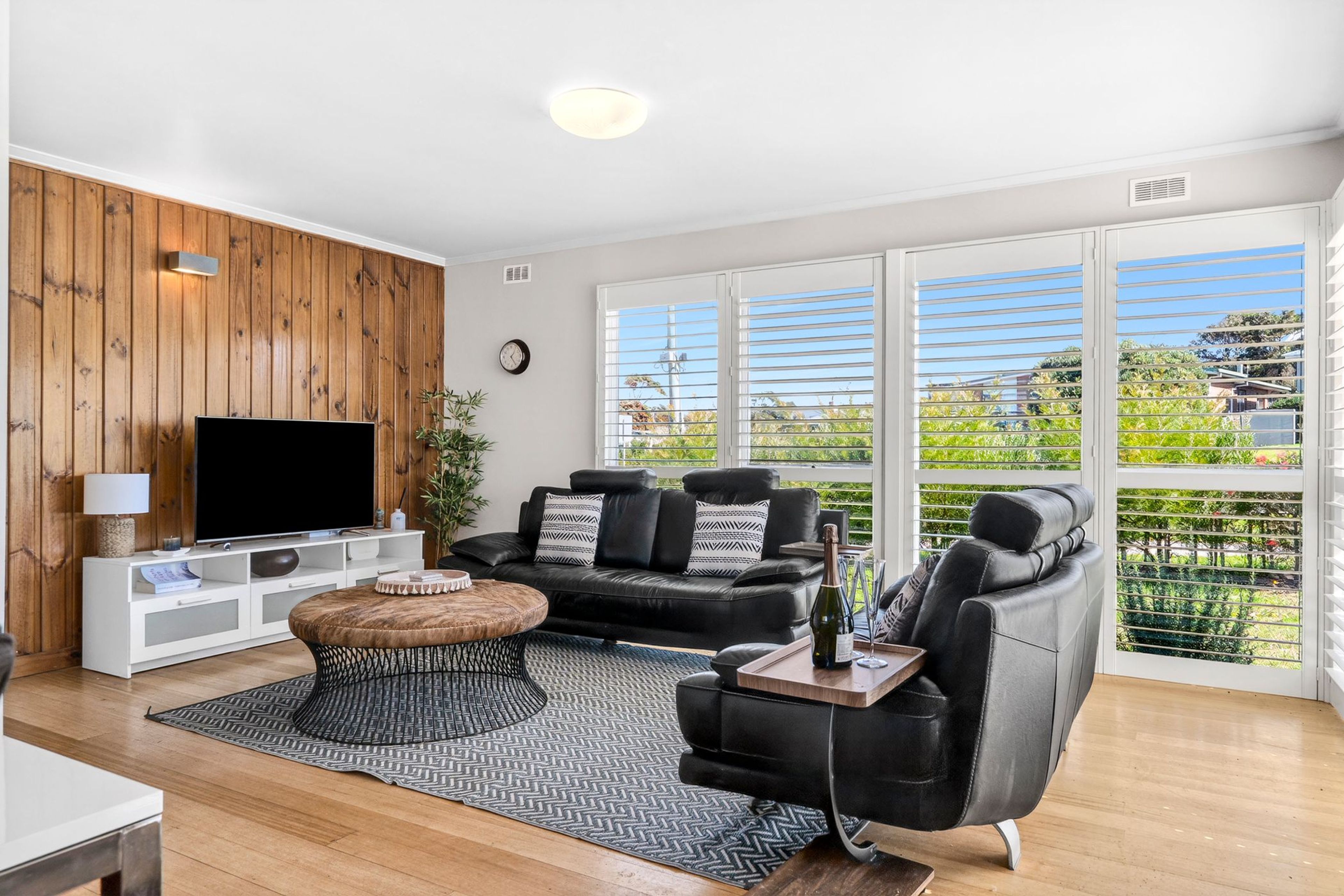 A modern living room with a flat-screen TV, a black leather sofa, a coffee table, and a large window with blinds.