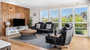 Een moderne woonkamer met een flatscreen televisie, een zwarte leren bank, een salontafel en een groot raam met zonwering.