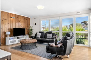 Ein modernes Wohnzimmer mit einem Flachbildfernseher, einem schwarzen Ledersofa, einem Couchtisch und einem großen Fenster mit Jalousien.