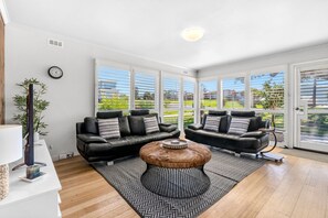 A modern living room with two black leather sofas, a round coffee table, and large windows offering a view of a garden.