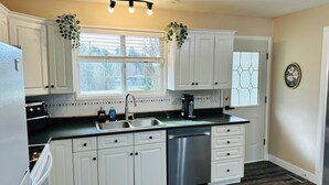A kitchen with white cabinets, a stainless steel dishwasher, a coffee maker, and a window with blinds. - Mainfloor 3 bedroom, sleep 5-6, Kelowna wineries retreats (Kelowna)