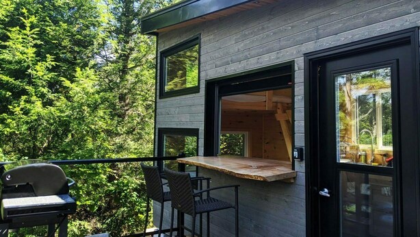 A modern cabin with a deck, built-in seating, and large windows overlooking a forest. - Baylight Cabin | Gorgeous Cabin on Private Lake (Dysart)