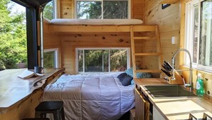 A compact cabin with a loft bedroom, a kitchenette, and a large window offering a view of the outdoors. - Baylight Cabin | Gorgeous Cabin on Private Lake (Dysart)