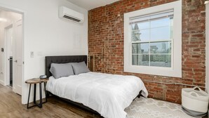 Una camera da letto con parete in mattoni, un'ampia finestra, un letto, un comodino e una lampada a sospensione.
