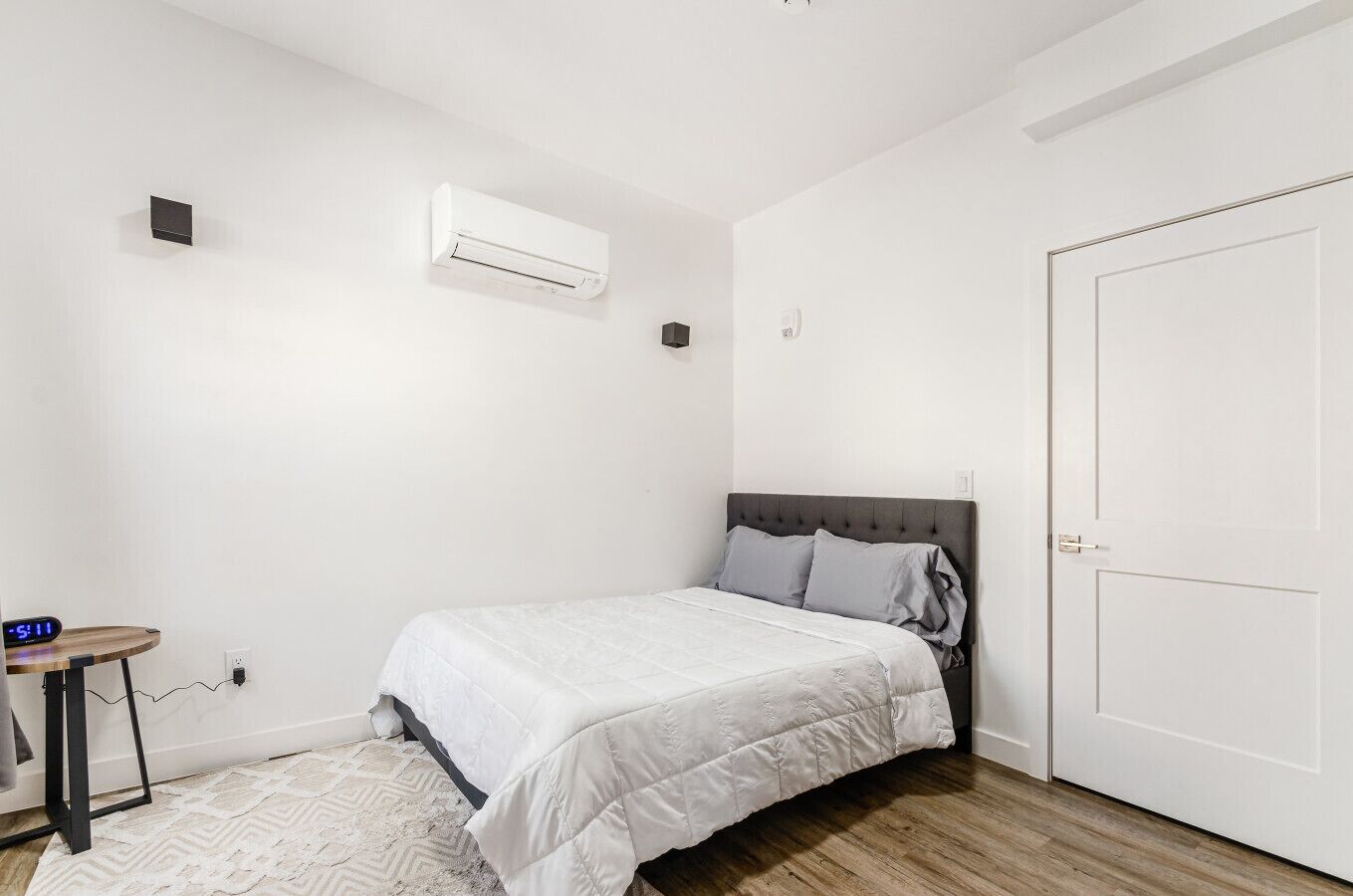 A bedroom with a bed, an air conditioner, a door, and a small table with a clock.