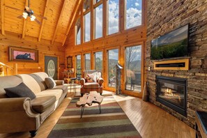 Interior acogedor de una cabaña con chimenea, ventanas amplias, un sofá y un sillón a cuadros.