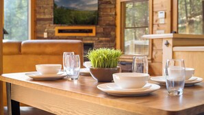 Un tavolo da pranzo in legno apparecchiato con piatti, ciotole e bicchieri bianchi, una pianta in vaso e un televisore montato su una parete in pietra.