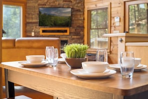 A wooden dining table set with white plates, bowls, and glasses, a potted plant, and a television mounted on a stone wall. - Smokies Getaway for 6 wPrivate Hot Tub & Mtn View (Sevierville)