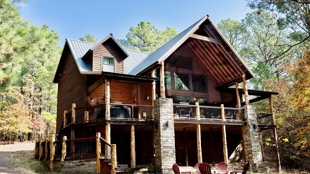Une cabane en rondins à deux étages, avec des fondations en pierre, des balcons en bois et une véranda équipée de fauteuils Adirondack.