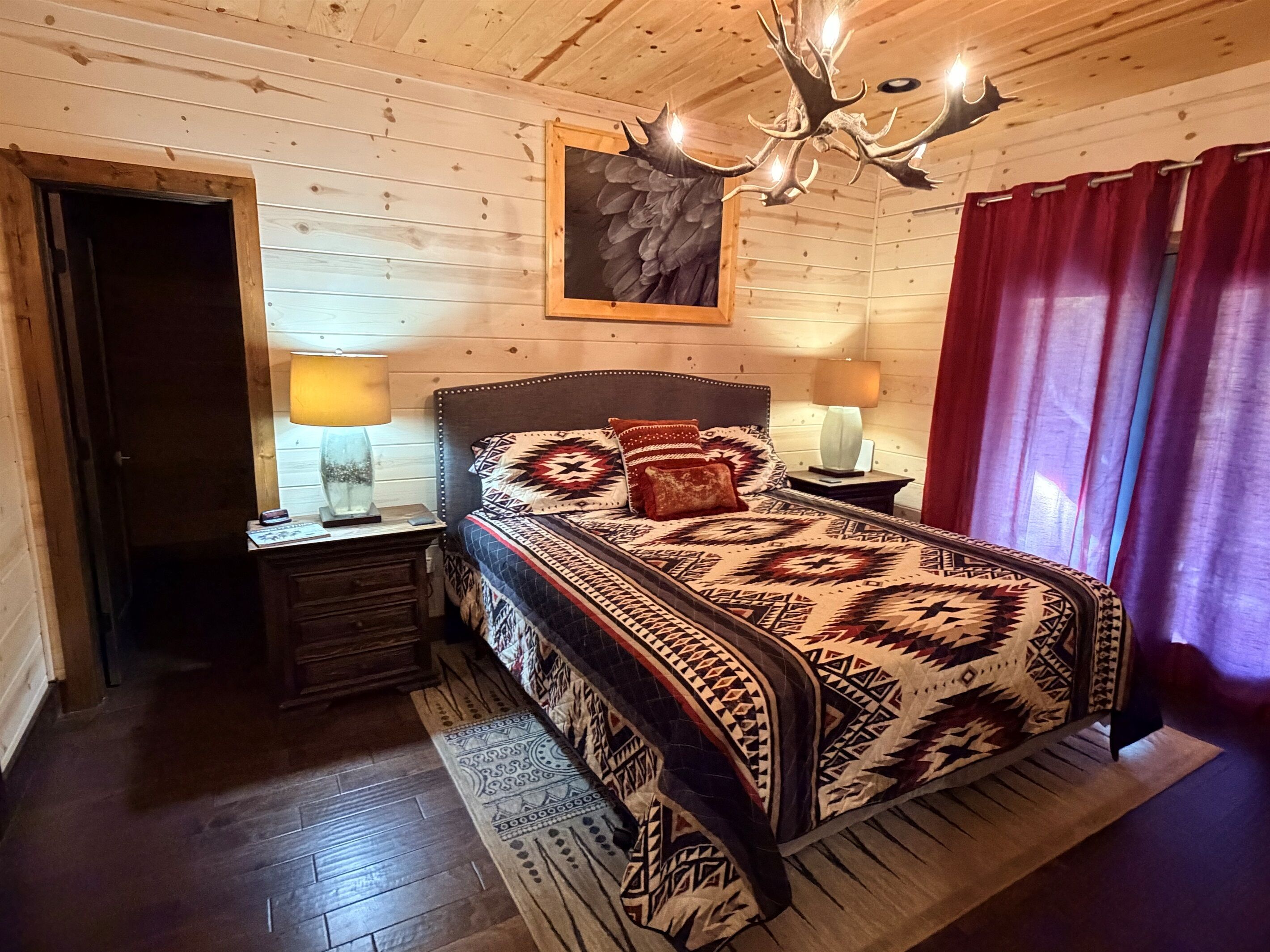 A wooden-paneled room with a bed, two bedside lamps, a chandelier, and a red curtain.