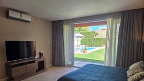 Quarto com cama, televisão sobre suporte de madeira, porta de vidro deslizante para um quintal e ventilador de teto.