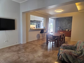 A modern living room with a flat-screen TV, a dining table with chairs, a patterned sofa, and a kitchen area with a countertop and cabinets.