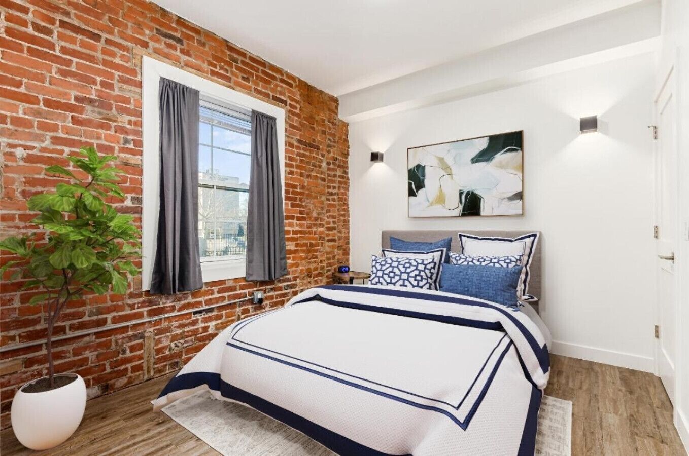 A bedroom with a brick wall, a large window with curtains, a bed with patterned bedding, a painting on the wall, and a potted plant.