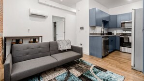 Un appartement moderne avec un canapé gris, un tapis à motifs, une cuisine aux meubles bleus et un appareil de climatisation.