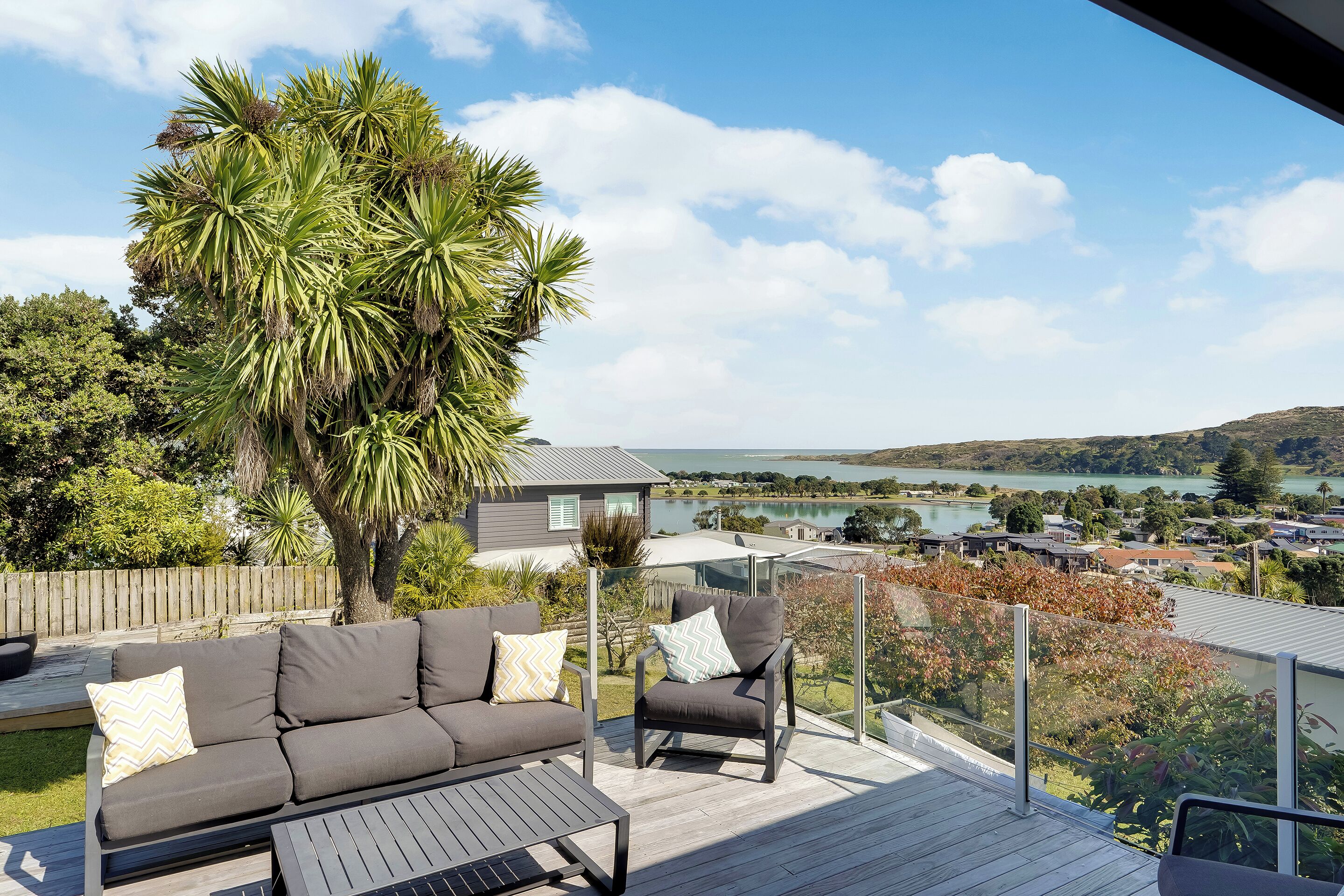 Une terrasse avec un canapé et un fauteuil, donnant sur un lac et des maisons.