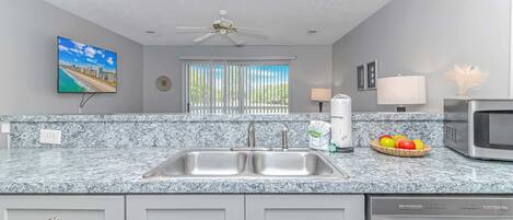 Una cocina con encimeras de granito, electrodomésticos de acero inoxidable, ventilador de techo y una ventana con persianas.