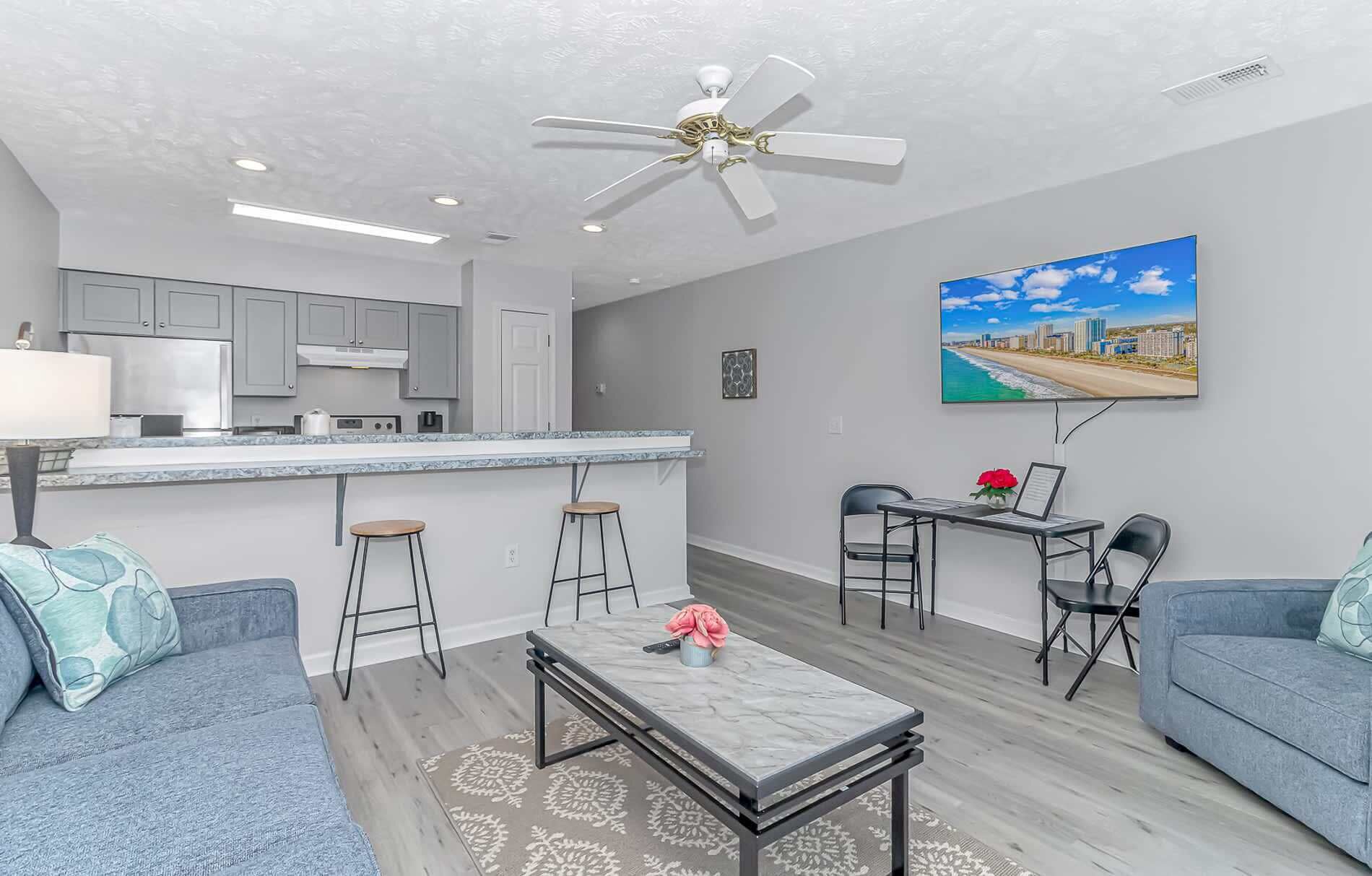 A modern living room with a grey sofa, a coffee table, a dining area with a table and chairs, a kitchen with grey cabinets, and a ceiling fan.