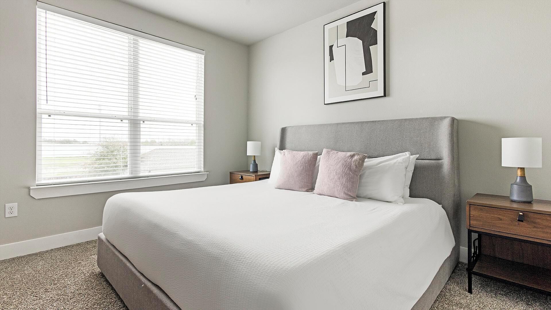 Een slaapkamer met een groot bed, een nachtkastje, een lamp, een raam met jaloezieën en een abstract kunstwerk aan de muur.