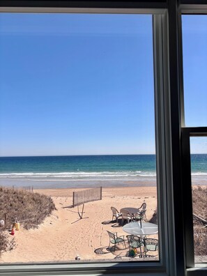 Blick durch ein Fenster auf einen Sandstrand, das Meer und einen Außenbereich mit Tischen und Stühlen.