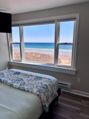 Ein Schlafzimmer mit großem Fenster mit Blick auf einen Strand, einem Bett mit floralem Überwurf und einem an der Wand montierten Fernseher.