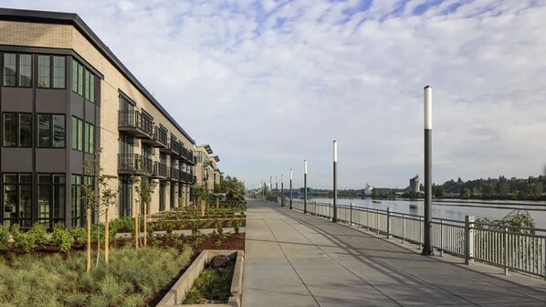 A modern building with large windows and balconies, a riverside walkway with metal railings, and a clear sky. - Landing | Incredible 1BD, Clubhouse (Portland)
