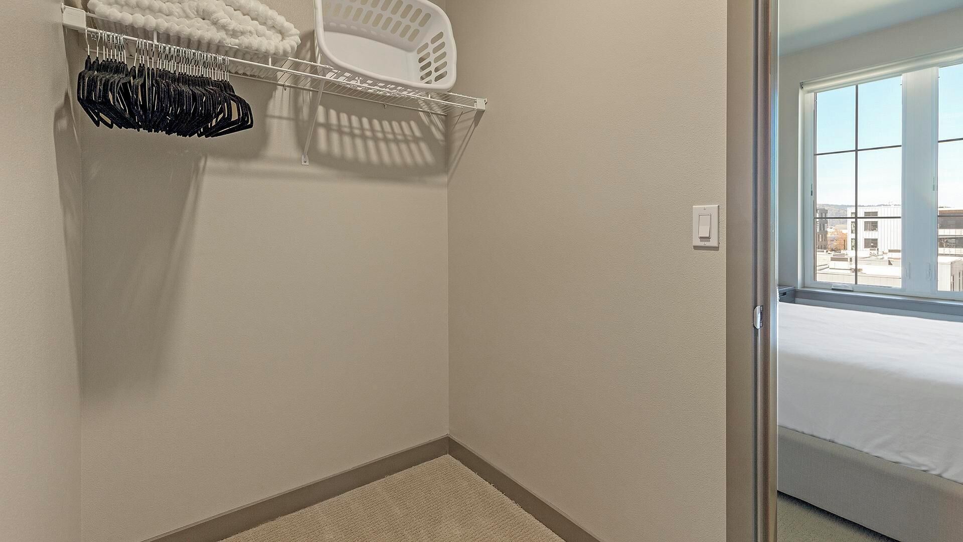 A corner of a room with a white closet, a white basket, and a window with a view of buildings.
