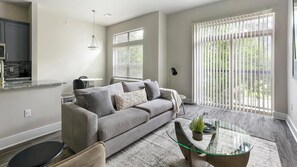 A modern living room with a grey sofa, a glass coffee table, and a view of the outdoors through large windows. - Landing | Spectacular 1BD, Yoga Studio, Clubhouse (League City)