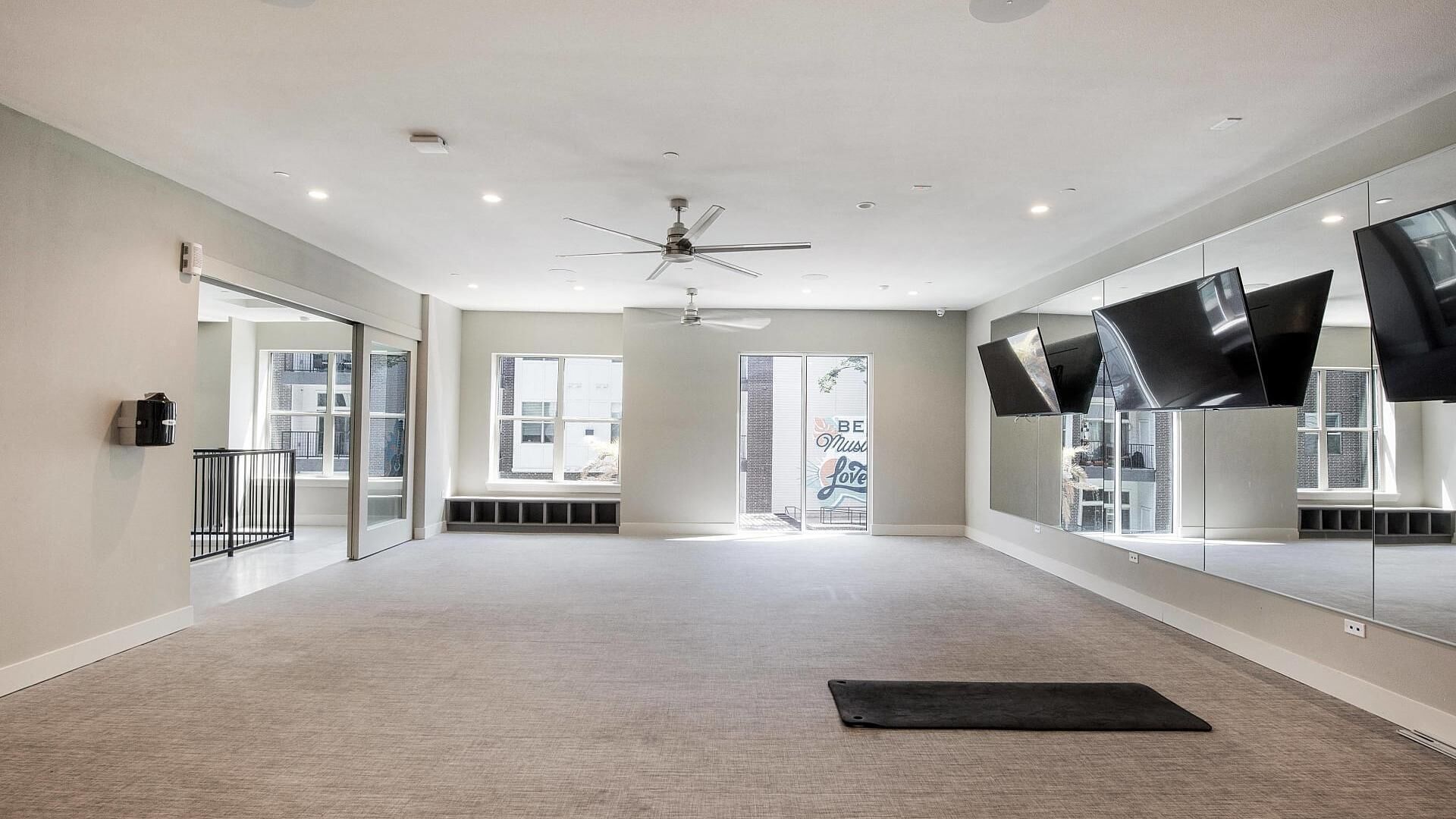 A spacious room with a ceiling fan, large windows, and a black mat on the floor.