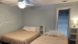 Une chambre avec deux lits, un ventilateur de plafond, une fenêtre avec des stores et une étagère fixée au mur, sur laquelle se trouve une lampe.