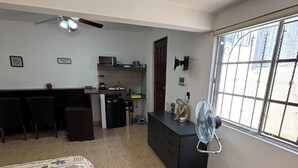 Ein Zimmer mit einem Bett, einem Schreibtisch, einer Küchenzeile, einem Standventilator und einem Fenster.