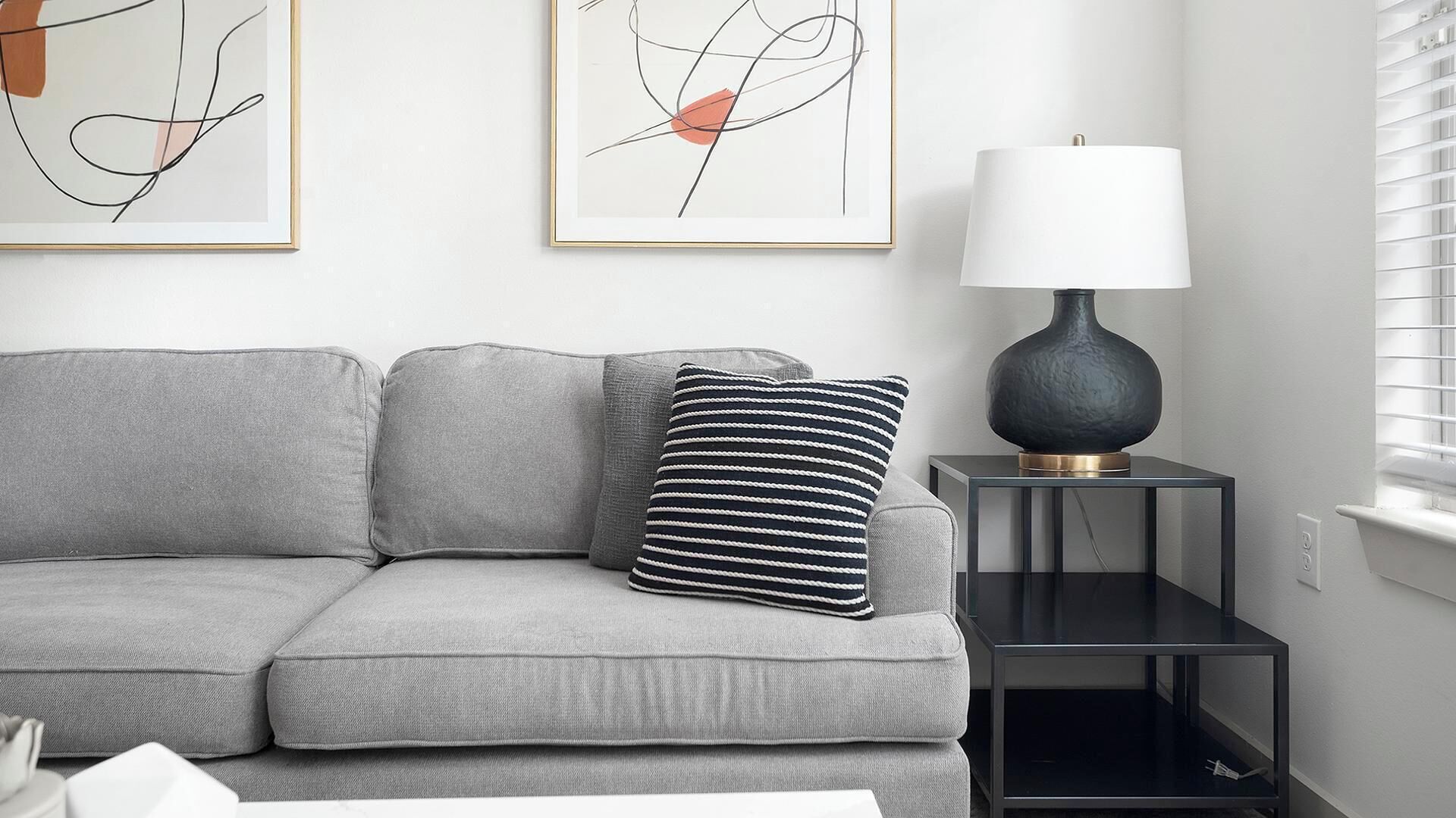 Ein modernes Wohnzimmer mit grauem Sofa, gestreiftem Kissen, abstrakter Wandkunst, einem schwarzen Beistelltisch mit Lampe und einem Fenster mit Jalousien.