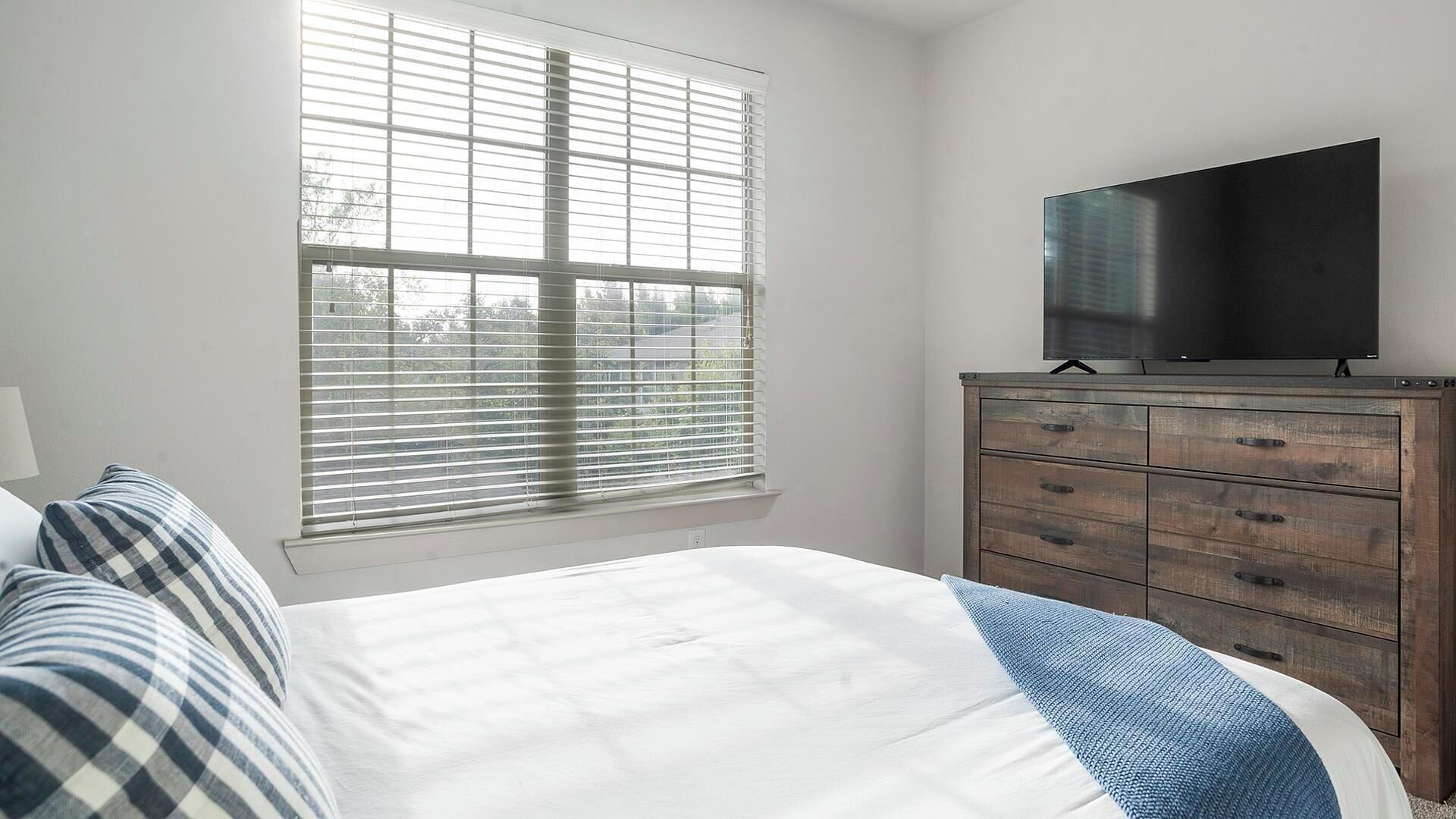 Une chambre à coucher comprenant un lit, une commode, un téléviseur et une fenêtre avec des stores.