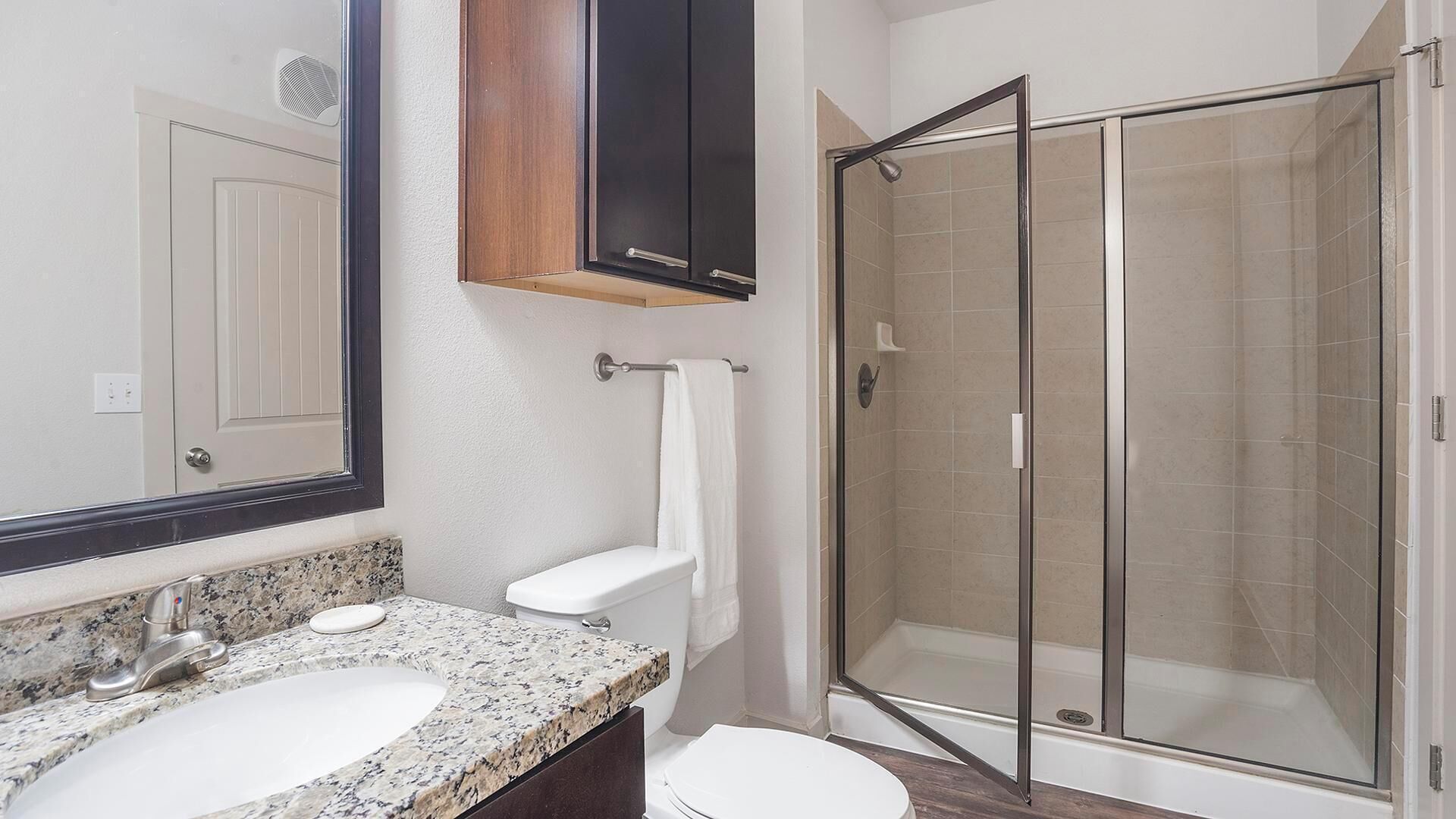 A bathroom with a granite countertop sink, a toilet, a glass-enclosed shower, and a mirror with a wooden cabinet above it.