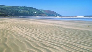 Une plage avec des ondulations dans le sable, une colline verdoyante à l’horizon et un ciel bleu clair.