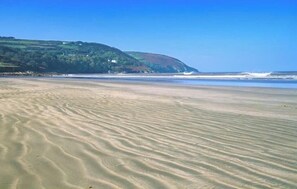 Ein Strand mit sanften Wellen im Sand, ein Hügel in der Ferne mit grüner Vegetation und einem strahlend blauen Himmel.