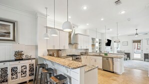 Cocina amplia con muebles blancos, encimeras de granito y electrodomésticos de acero inoxidable. Incluye tres taburetes de bar y lámparas colgantes.