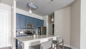 A modern kitchen with blue cabinets, a stainless steel microwave, a refrigerator, and a granite countertop. There are two bar stools and a pendant light above the island. - Landing | Stylish 2BD, Clubhouse, Pool (League City)