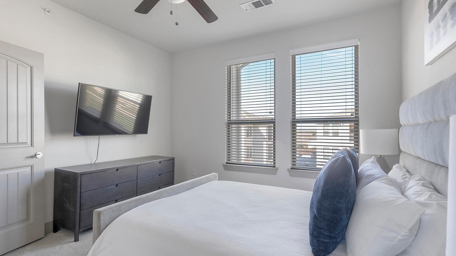 A bedroom with a bed, a dresser, a TV, and two windows with blinds.