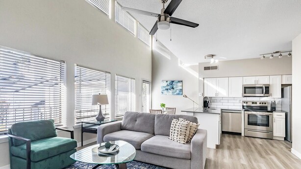 A modern living room with a sofa, a glass coffee table, and a green armchair. There is a kitchen with stainless steel appliances and a ceiling fan. - Landing | Incredible 1BD, Pool, Gym (Fort Worth)
