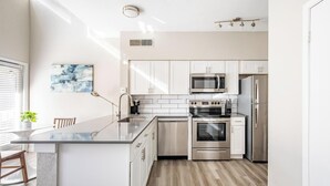 A modern kitchen with stainless steel appliances, a central island, and a view of the dining area. - Landing | Incredible 1BD, Pool, Gym (Fort Worth)