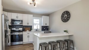 Uma cozinha moderna com armários brancos, eletrodomésticos de aço inoxidável, uma ilha grande com banquetas e um relógio de parede.