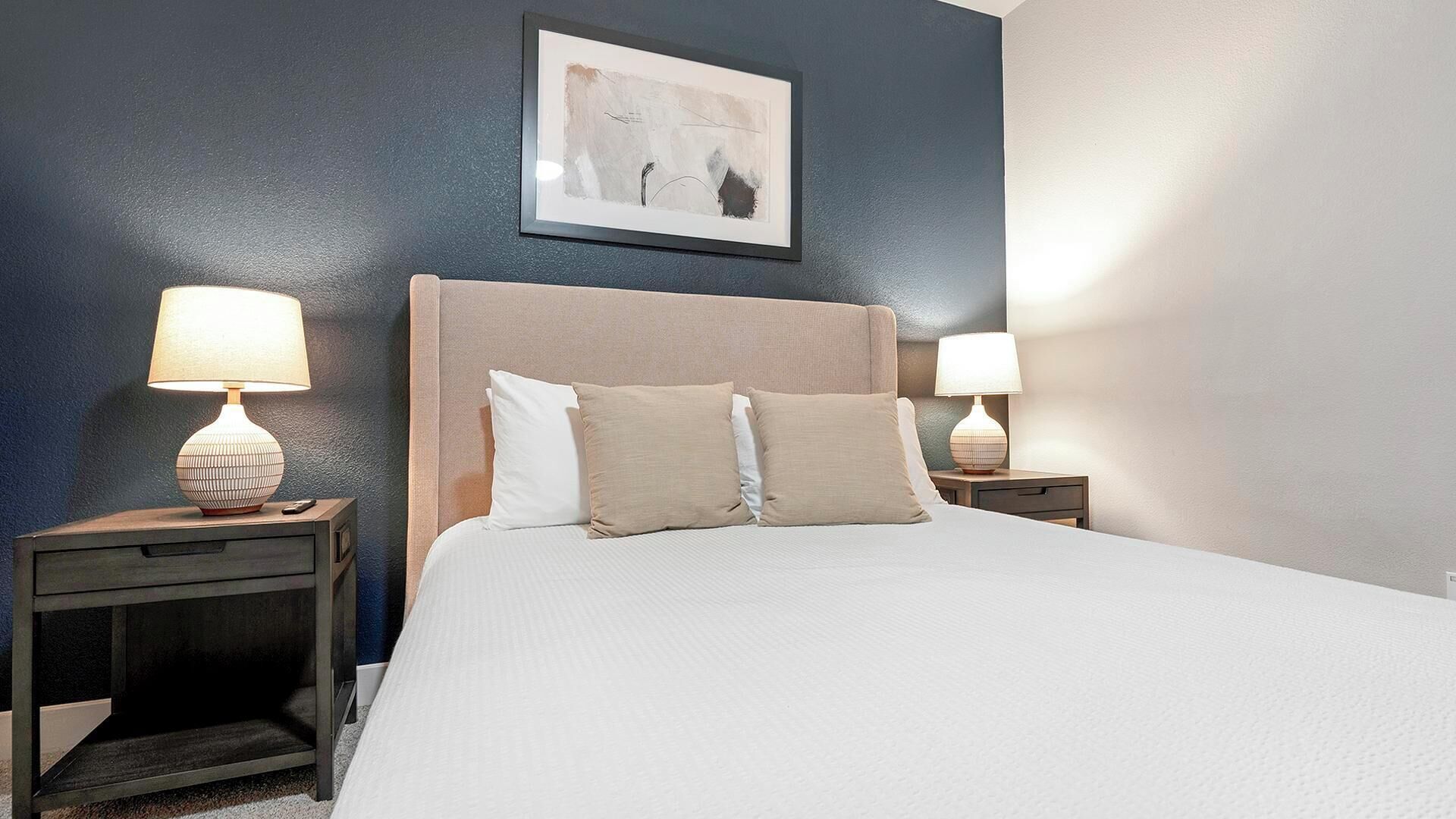 Et hotelværelse med en seng, to sengeborde med lamper, et abstrakt kunstværk i ramme på væggen og en mørkeblå accentvæg.