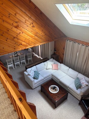 Ein Loft-Wohnzimmer mit Holzbalkendecke, einem weißen Ecksofa, einem Couchtisch und einem Essbereich mit Stühlen und Tisch.