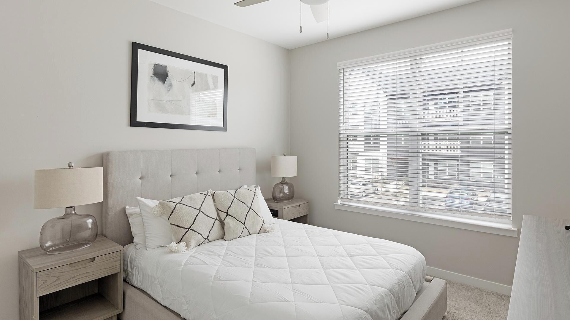 Une chambre à coucher avec un lit, deux tables de chevet avec des lampes, un ventilateur de plafond, une fenêtre avec des stores et un tableau encadré au mur.