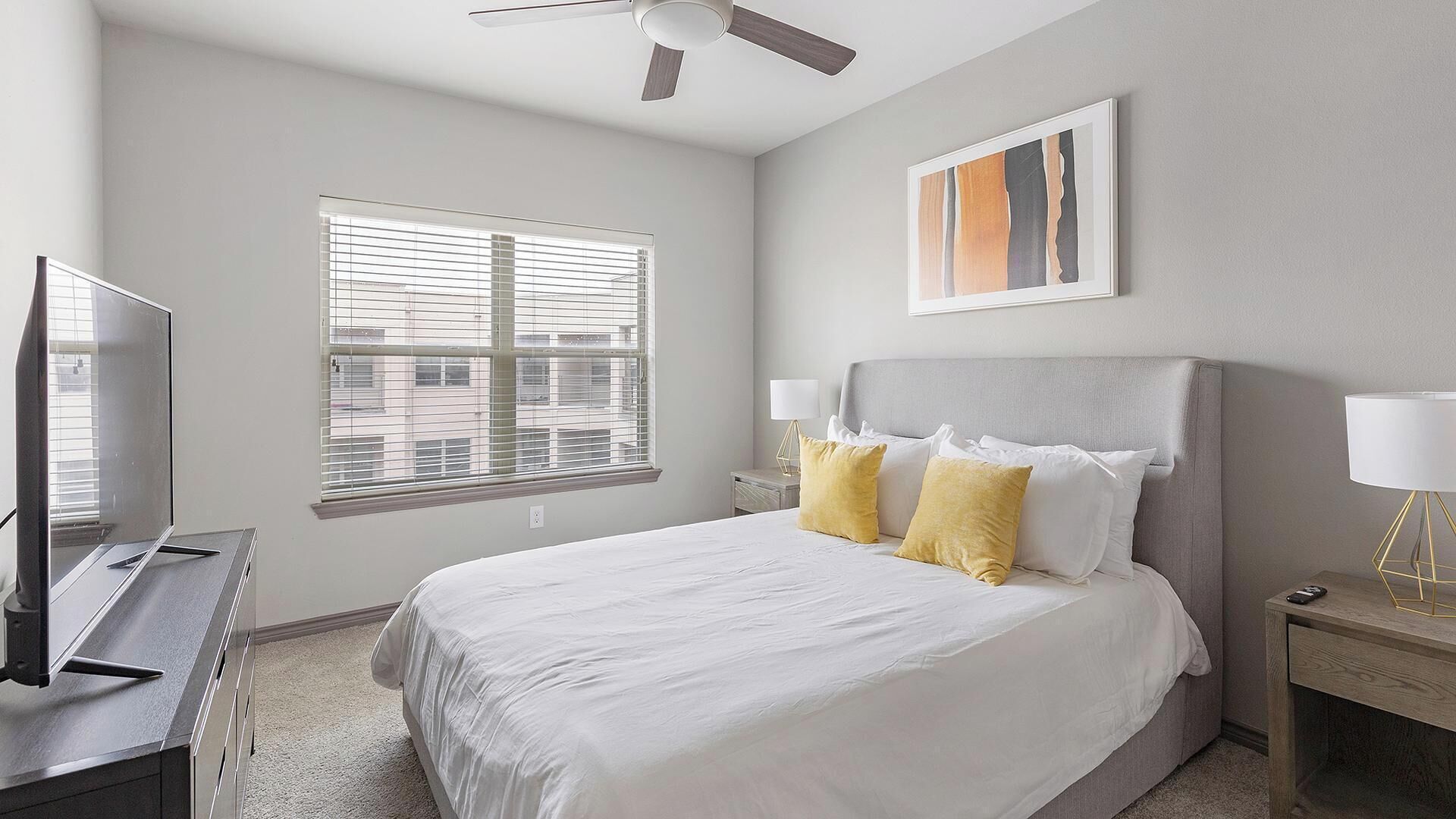 Une chambre à coucher avec un lit, un téléviseur, un ventilateur de plafond, une fenêtre avec des stores et un tableau au mur.