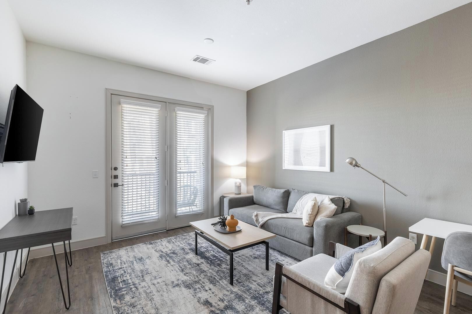 A modern living room with a grey sofa, a coffee table, a TV mounted on the wall, and a large window with blinds.