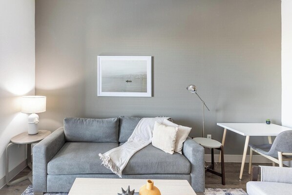 A living room with a grey sofa, a small round table, a white coffee table, and a framed picture on the wall. - Landing | Spectacular 1BD, Clubhouse, Gym (Denton)