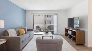 A modern living room with a sofa, a coffee table, a TV stand, and a sliding glass door leading to an outdoor area. - Landing | Chic 2BD, Courtyard (Paradise)